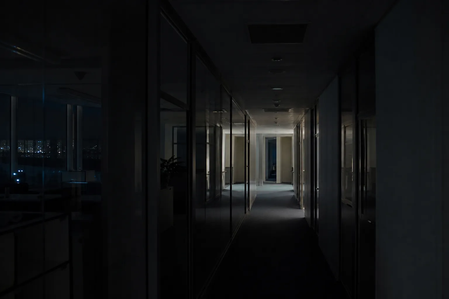 Couloir de bureaux éclairé de nuit, une seule zone allumée, ville sombre derrière les baies